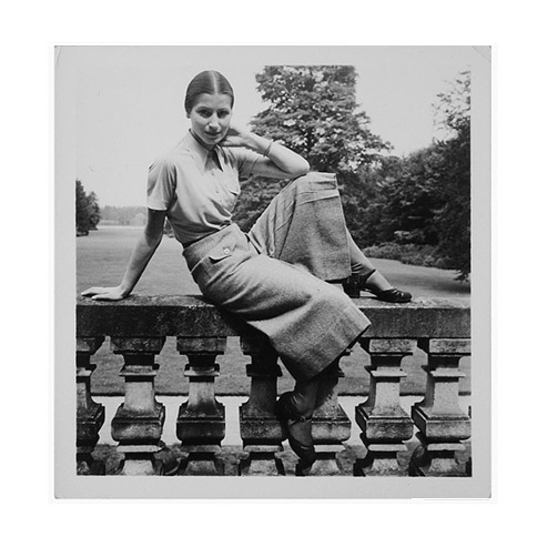 Porträtfoto einer jungen Frau im Hosenrock, die anmutig auf einer alten Brüstung posiert. Hinter ihr erstreckt sich ein großer Garten.