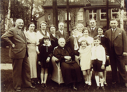 Gruppenfoto mit neun Erwachsenen und sechs Kindern. In der Mitte eine ältere Dame in einem Korbsessel. Das Foto ist in einem Garten aufgenommen, im Hintergrund sind ein großes Haus und Bäume zu sehen