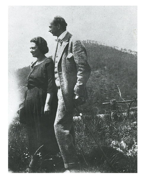 Paar beim Spaziergang. Die Frau in einem dunklen Sommerkleid, der hochgewachsene Mann in Anzug und mit einem Spazierstock. Um sie herum Gras, im Hintergrund ein Hügel.
