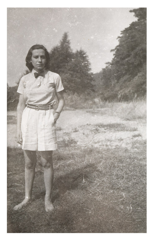 Junge Frau in sommerlicher kurzer heller Hose und Bluse mit dunkler Fliege. Sie steht barfüßig auf einer Wiese mit Sandflecken, im Hintergrund sind Bäume zu sehen.
