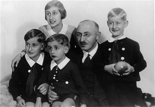 Gruppenfoto eines erwachsenen Mannes mit 3 Jungs und einem Mädchen. Die Kinder sind fein angezogen und sorgfältig gekämmt, der Mann trägt dunklen Anzug und Krawatte. Sie blicken fröhlich oder etwas verlegen.