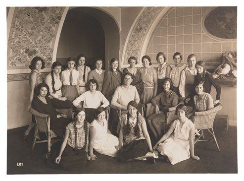 20 junge Frauen in sommerlicher Kleidung, die sich zu einem Klassenfoto aufgestellt haben, in der Mitte sitzt ihre Lehrerin.