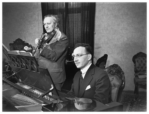 Schwarzweißfotografie, die zwei Männer beim Musizieren in einem bürgerlichen Ambiente zeigt. Der jüngere mit Brille und Anzug sitzt am offenen Flügel, der ältere, mit auffallend weißem lockigem Haar steht daneben und spielt die Violine.