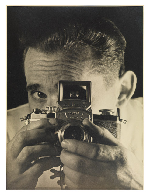 Photograph showing a man with his face concealed behind a camera that he holds in both hands; one of his eyes is visible through the viewfinder.