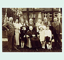 Group photo of nine adults and six children with an elderly lady sitting in the middle. The photo was taken in the garden and a large house and trees can be seen in the background.