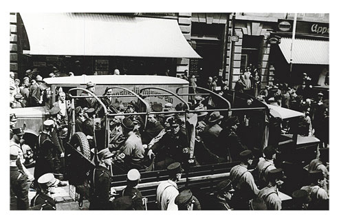 An open truck on the bed of which men sit crowded together on benches, some in uniform. The truck is driving down a street, accompanied by uniformed marchers. Passersby look on from the sidewalk.