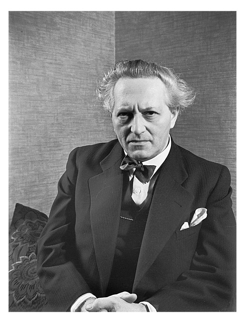 Man sitting in a dark suit with a bow tie and a handkerchief in his breast pocket. His hands are folded in his lap and he has a serious expression on his face.