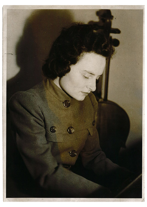 Portrait photo of a dark-haired young woman, seated, in a jacket with large buttons. The position of the hands suggests that she is playing the piano, but we cannot see the instrument. There is a cello in the background.