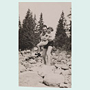 Vertical-format photo showing a man in a bathing suit with a girl in his arms wearing a summer dress. They are standing amidst large stones, perhaps in a dry river bed, and looking cheerfully at the camera. Fir trees can be seen in the background.