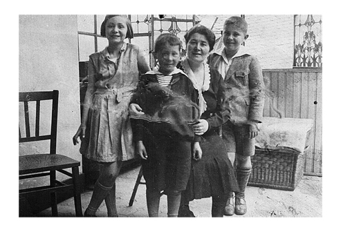 Black-and-white photo of a woman with a girl and two boys in a conservatory. The woman is seated on a chair with the children standing around her smiling at the camera.