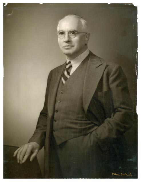 Photo portrait of an elderly man in a suit and glasses with combed-back white hair.