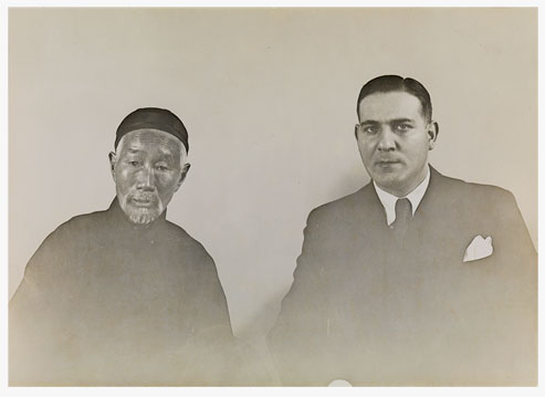 An elderly Asian man in a traditional Chinese robe and hat, next to him a young man with European features in a Western suit, his hair neatly parted.