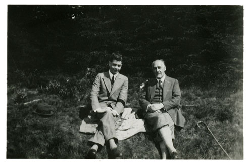 Two men sit on the grass on a rug or coat. Beside them are a hat and a hiking stick. The young man on the left is wearing a suit and tie, and smiles rather shyly into the camera. The man on the right is considerably older, bearded and balding, and wears knickerbockers with a jacket and vest.