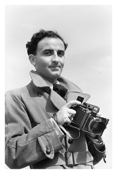 Man in a trench coat holding a large camera.