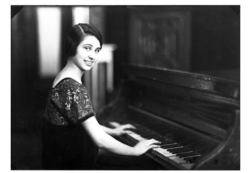 A young woman with short dark hair sitting at a piano smiling at the camera.