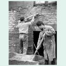 Two young men in their twenties or teens working in front of a brick wall. The young man on the right, dressed in a work coat, is mixing plaster with a stick in a wooden tub. The one on the left, wearing a shirt, pants and cap, is spreading the mixture on the wall with the help of a large board with a handle.