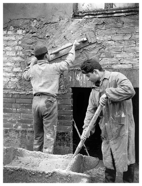 Two young men in their twenties or teens working in front of a brick wall. The young man on the right, dressed in a work coat, is mixing plaster with a stick in a wooden tub. The one on the left, wearing a shirt, pants and cap, is spreading the mixture on the wall with the help of a large board with a handle.