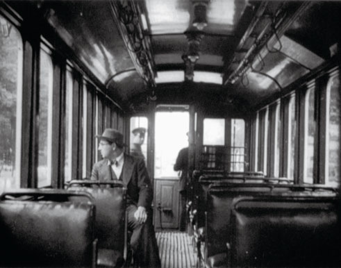 Man with a hat looking out the window of a train compartment, with a conductor in the background (historical image)