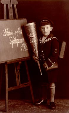 Boy in a sailor's suit with a school bag in front of a chalkboard reading My first day of school 193...