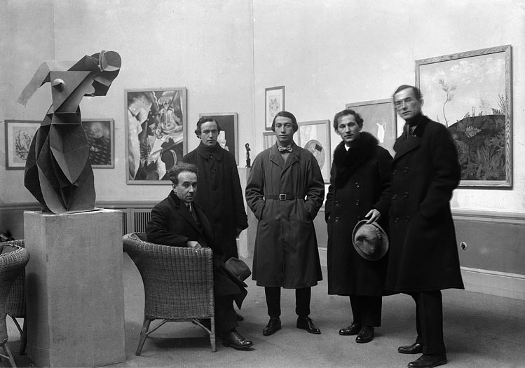 5 Männer in Mänteln in einem Ausstellungsraum, im Hintergrund Gemälde, links eine Skulptur