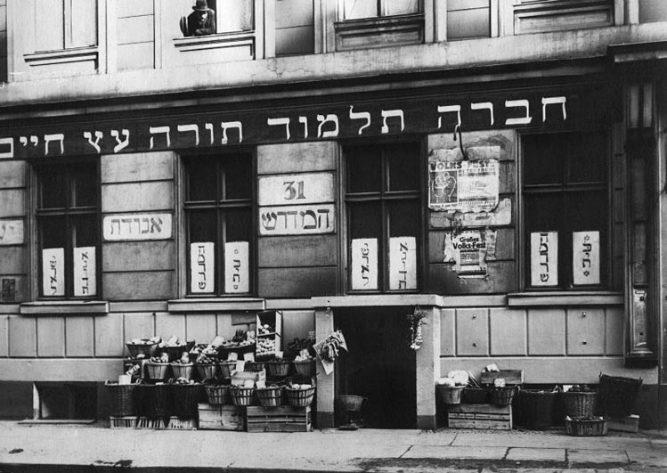Schwarz-weiß Foto einer Hausfassade mit Körben vor der Tür und Schildern (hebräische Schrift)