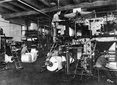 View of a room with rotary printing presses; one worker is standing on a ladder and others can be seen in the background