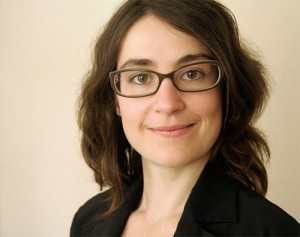 A woman with glasses, black clothes and long brown hair looks directly at the camera