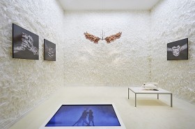 A white room with feathers, black and white photos of hands, a sculpture of hands and a swan
