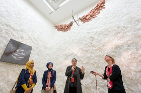 Four women in a white room with feathers on the wall, from the floor ins hanging an artwork: wings made of human hands