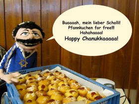 A puppet in a blue shirt with the star of David, in front of a crate of Berliner hotcakes with a speech bubble, “Oooh, my oh my! Hotcakes for free!!! Hahahaaaa! Happy Hanukkaaaah!”