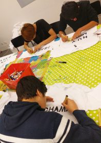 Three boys sitting around a table and writing their names on T-shirts