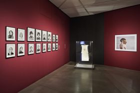 View of a room in the exhibition with portraits of women on one wall, a dress in a glass case, and a photo of a woman with two wigs on another wall.