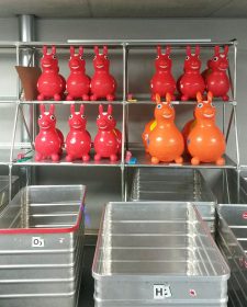 Red rubber bouncy animals that resemble horses on a gray shelf behind empty wheeled containers