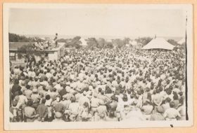 On the black and white picture photographed from diagonally above, a large crowd of people faces a house with a man standing on its roof and speaking to the crowd.
