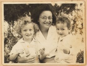 Black and white photography: Leonie is sitting in the middle and laughs. Michael, who runs his tongue over the right corner of his mouth, is sitting on her lap.On the left is Peter-Uri with bright curls, also smiling broadly.