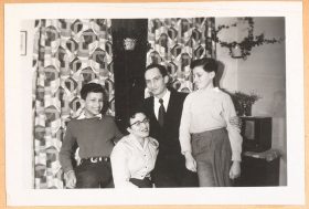 In the black-and-white photo, the family is in a room with patterned curtains and houseplants. All four are smiling or laughing. The image is very lively.