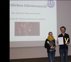 Under a screen on which the Traineeship Committee congratulates the winners of the prize, Franziska and David hold the certificate up to the camera.