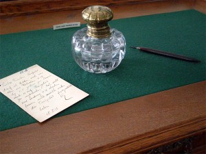 Gläsernes Tintenfass, Schreibfeder und Postkarte auf einem Schreibtisch