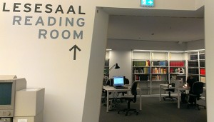 Im Vordergrund eine Wand mit der Aufschrift »Lesesaal«, im Hintergrund Bücherregale und Lesetische