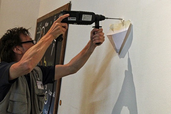 Ein Mann hält eine Bohrmaschine an die Wand, unter dem Bohrloch hängt eine gefaltete Papiertüte