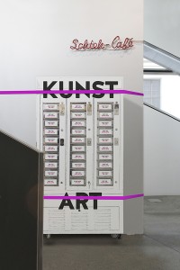 Ein weiß lackierter Warenautomat mit der Aufschrift »Kunst / Art«, mit pinken Bändern an eine Wand mit der Aufschrift »Schteh-Café« gebunden