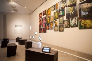 44 Portraits hängen an der Wand, davor schwarze Bänke, auf denen iPads liegen