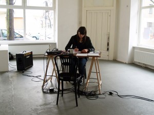 Foto der Künstlerin, sie sitzt an einem Schreibtisch in in einem ansonsten fast leeren Raum