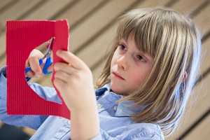 Ein Mädchen schneidet mit einer Schere etwas aus einem roten Karton aus.