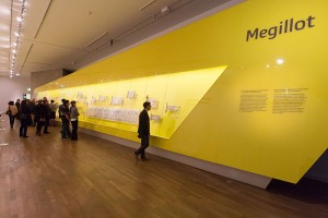 Personen stehen in einem gelb gestrichenen Raum vor einer sehr langen Galsvitrinen, die auf der rechten Seite in eine gelbe Ausstellungsfläche eingearbeitet wurde. In der Vitrine hängen verschiedenen Schriftrollen.