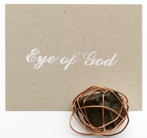 Ein mit draht umwickelter Schotterstein liegt neben einer grauen Karte auf der mit weißer Schrift "Eye of God" steht
