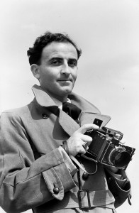 Schwarzweißaufnahme eines Mannes der eine Kamera in den Händne hält und direkt den Fotografen des Bildes anschaut