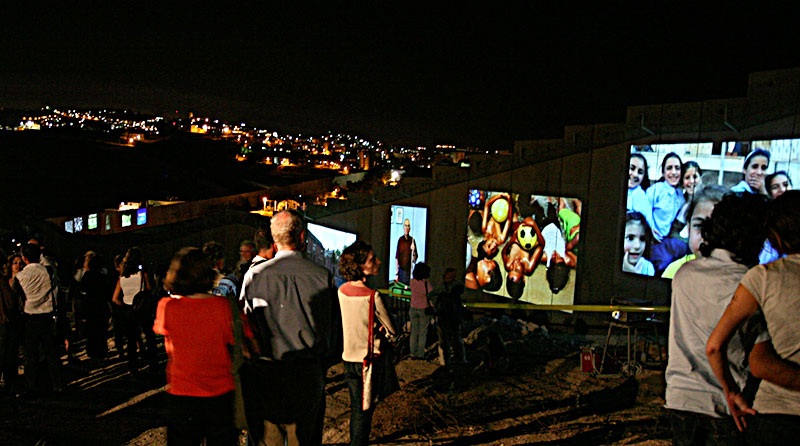 Farbfotografie einer Kunstinstallation mit Bildprojektionen an eine Mauer im Dunkel mit Menschen davor und einem Weitblick auf dahinter liegende beleuchtete Wohnhäuser