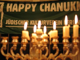 Chanukkaleuchter mit brennenden Kerzen, dahinter ein Banner mit der Aufschrift "Happy Chanukka. Jüdischer Kulturverein Berlin"