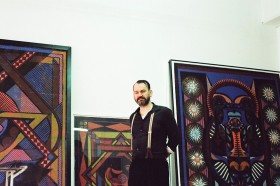 Ein schwarz gekleideter Mann mit Hosenträgern steht zwischen großformatigen Bildern